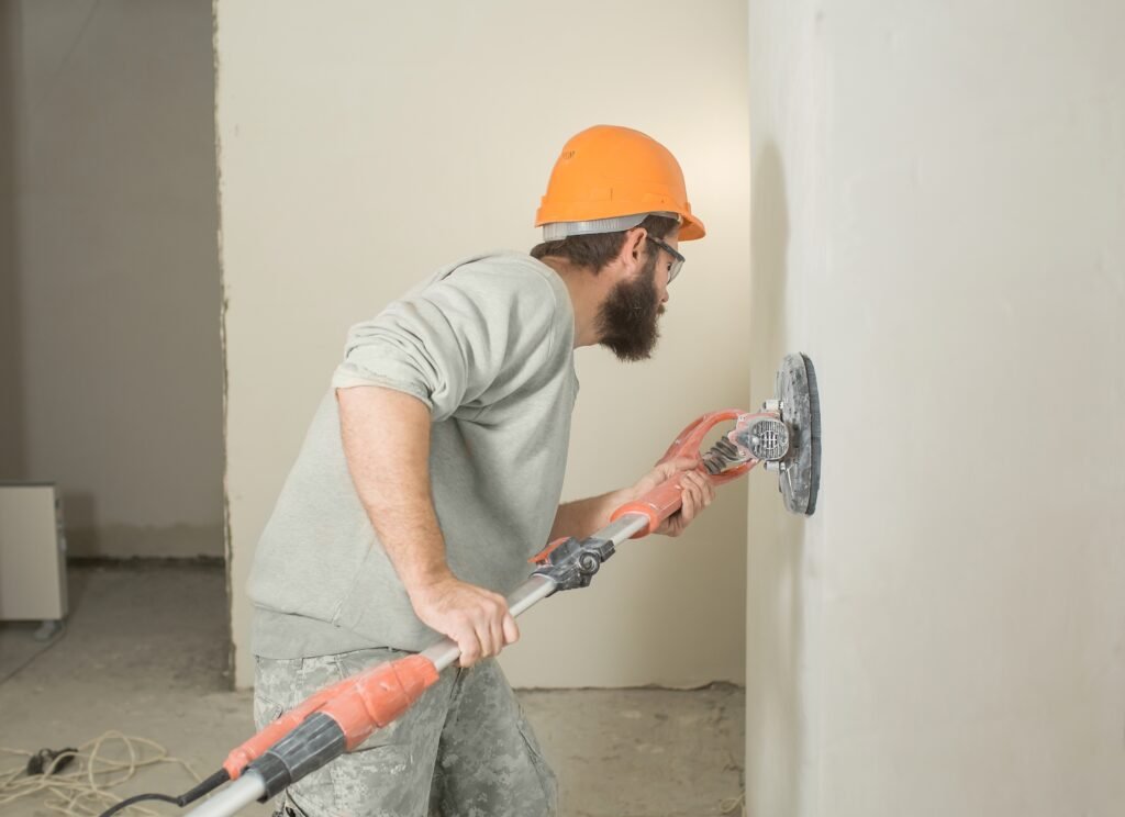 a man with a beard in a construction helmet skins the walls with an electric grinder