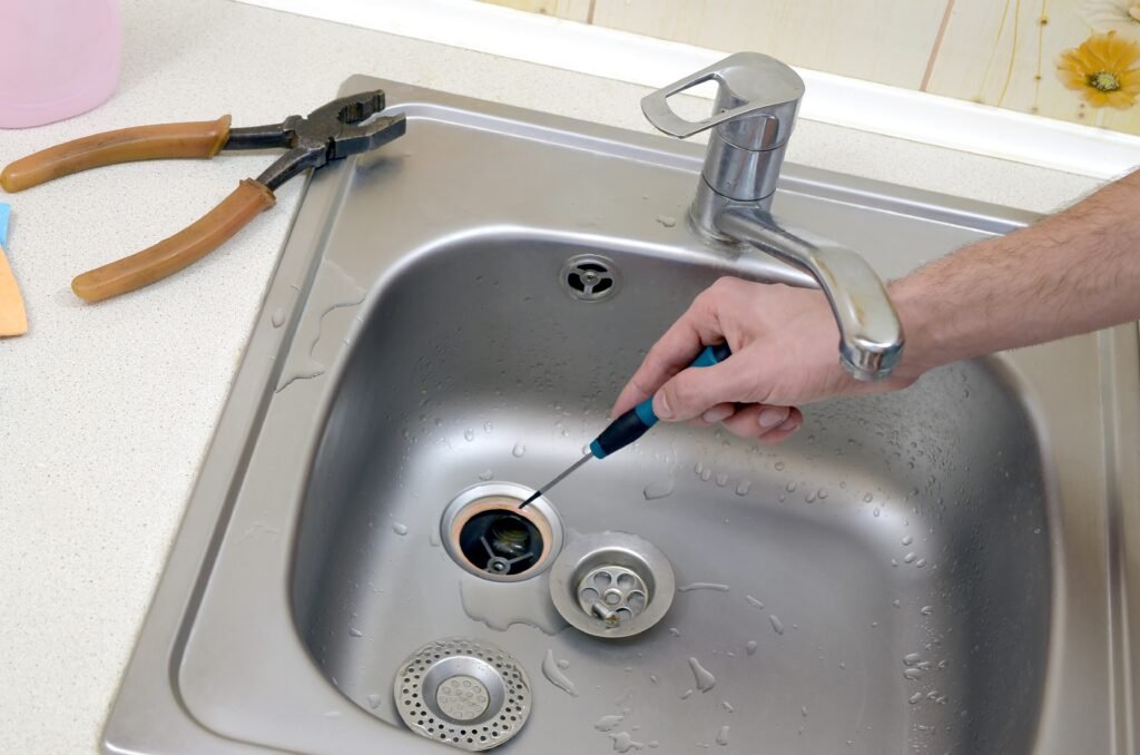Close-up of handsome plumber repairing drain of kitchen sink. Photo of metalic sink and handyman with screwdriver