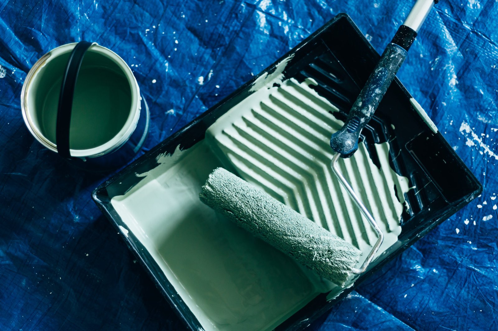 Painting and refreshing the wall with green paint, brushes, roller, tray and can of paint. Floor is protected with blue cover. Premium Interior Wall, Ceiling and Wood Paint