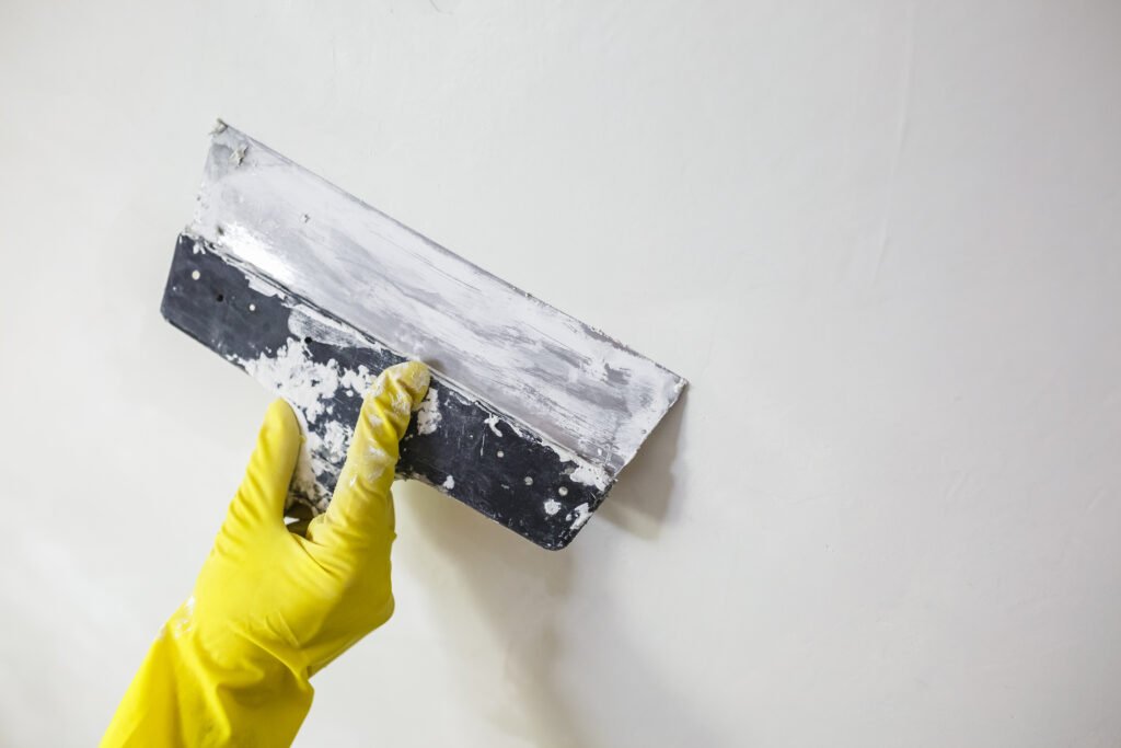 worker's hand in yellow gloves holding putty knife patching a hole with spatula with plaster or putty in white wall. Renovation and repair process, remodeling interior of room at apartment building